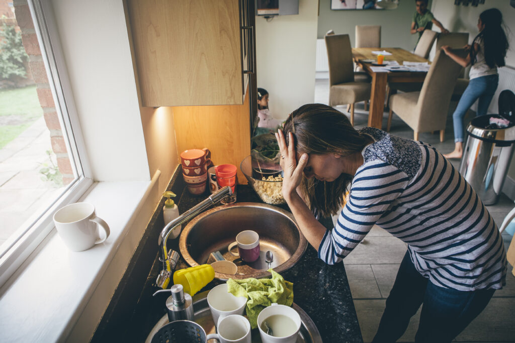 Stressed mom at home. She has her head in her hands at a messy kitchen sink and her children are running around in the background.