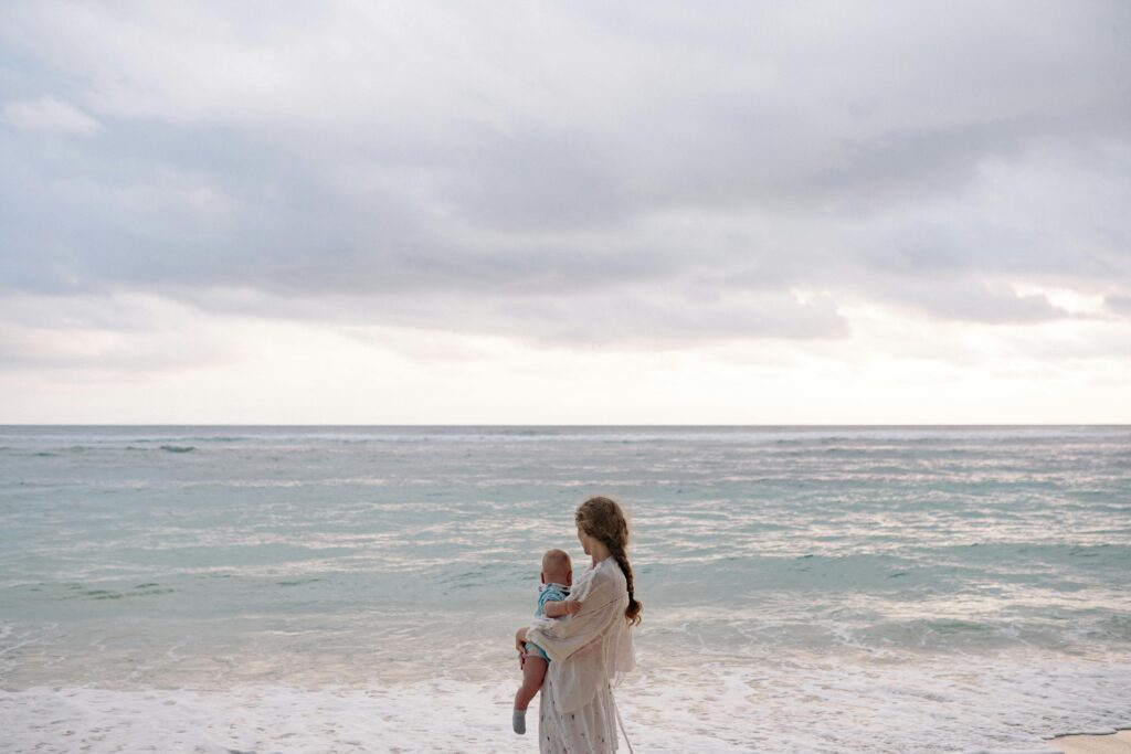 A single woman holding baby by the ocean and looking out at the water